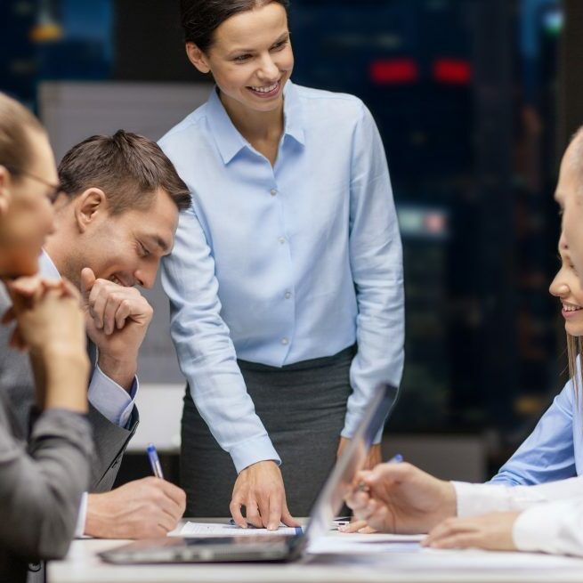 business, technology, people, deadline and team work concept - smiling female boss talking to business group at night office background